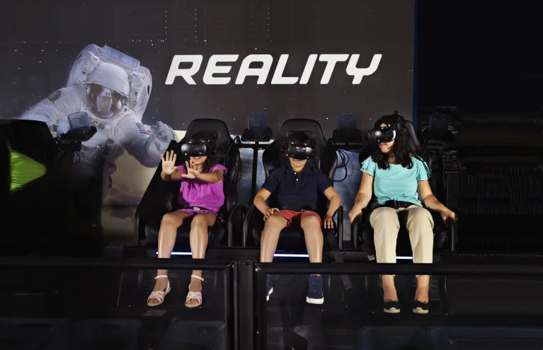 Image of a boy, woman and a girl with arm stretch out as they ride the Apollo 11 & Beyond VR Experience
