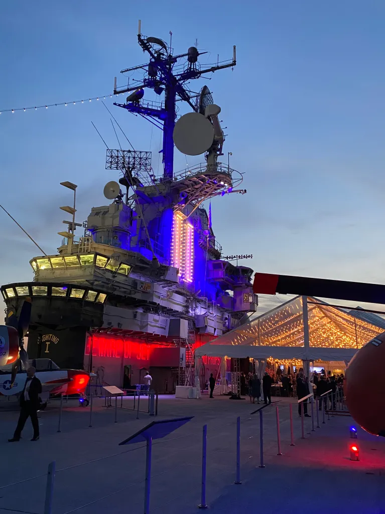 The Center of the Flight Deck is tented with a clear tent for an evening event with string lights and uplighting on the Flight Deck Island